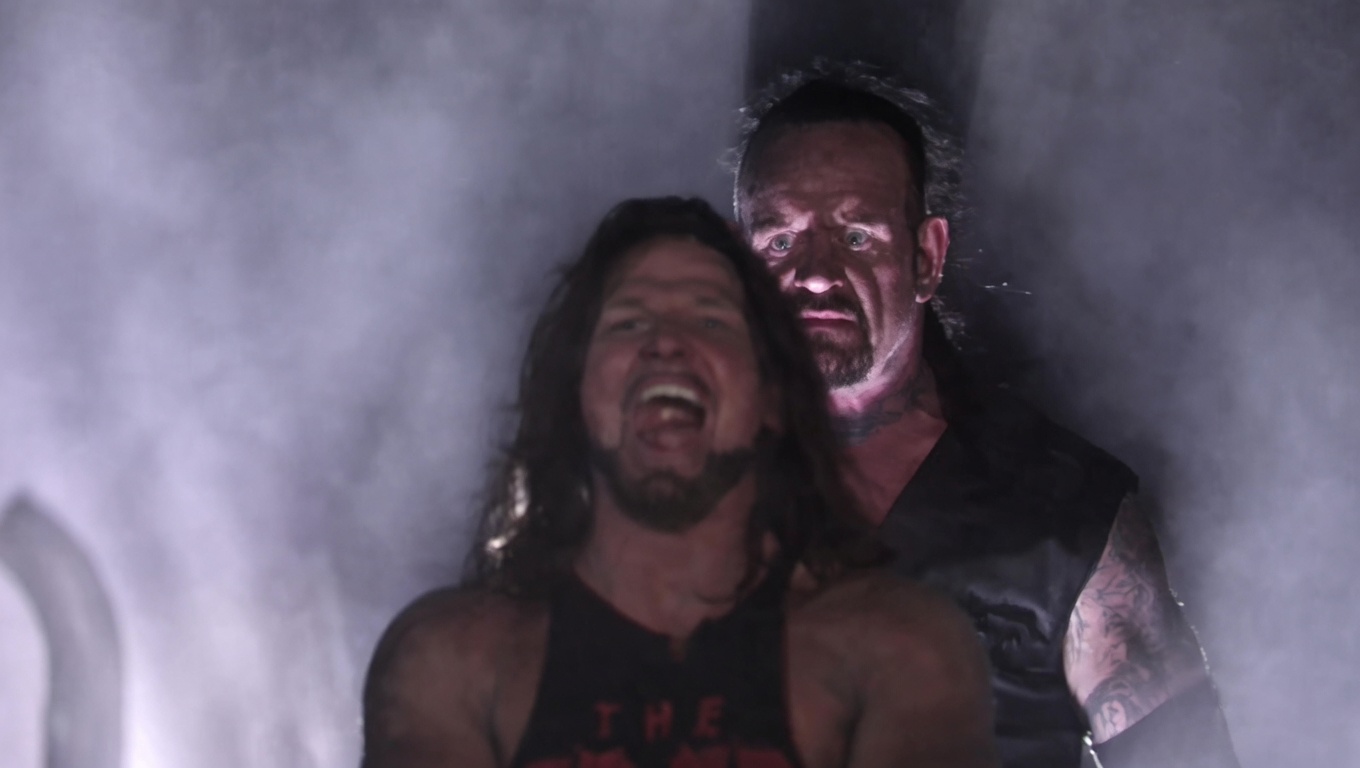 Two men stand in thick gray smoke under dim, dramatic light. In front, a long-haired, bearded man in a black tank top with partial red lettering has his mouth wide open in a scream. Behind him and slightly higher, a stern man with slicked-back hair, a goatee, neck and arm tattoos, and a black sleeveless vest stares forward calmly.