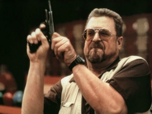 A middle-aged man with a goatee, mustache, and tinted glasses wears a dark shirt under a light vest and a black watch. He holds a handgun raised in both hands and stares forward with a stern, intense expression. Bowling balls and alley seating are visible behind him.