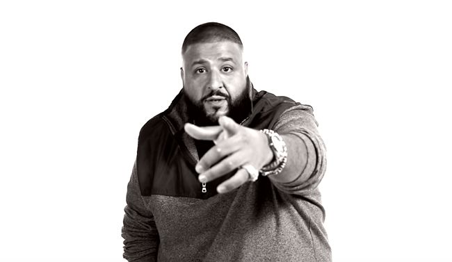 A black-and-white studio photo of a bearded man with a short haircut, wearing a zip-up hoodie with darker shoulder panels. He faces the camera and points straight at the viewer, one hand extended forward and the other also raised. A watch and rings are visible. His mouth is slightly open. The background is plain white.