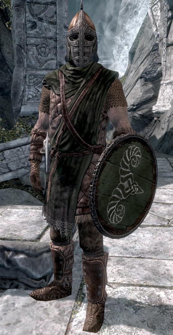 A full-body armored guard stands facing forward on pale stone pavement. He wears a metal helmet with a pointed crest and a barred face guard, a dark green quilted tunic over chainmail, brown leather straps, gloves, and tall studded boots. A round green shield with a metal rim and a white curling emblem is held at his side, and a sword hilt shows at his hip. Behind him are a carved stone doorway, rocky cliffs, and a waterfall.