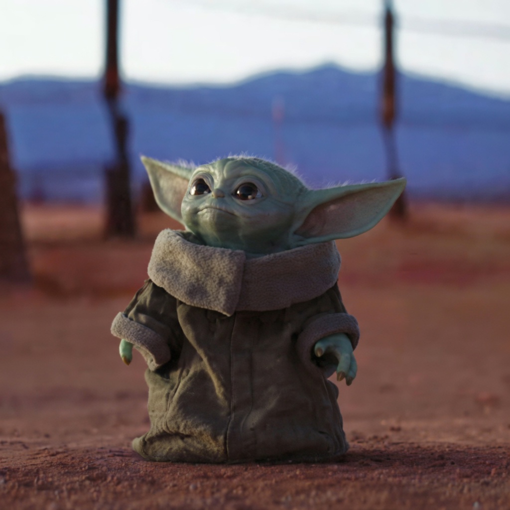 A small green creature with huge pointed ears, large dark eyes, and a slight closed-mouth smile stands centered on reddish dirt. It wears an oversized brown robe with a thick beige fleece collar and cuffs, and its three-fingered hands hang at its sides. The background is a soft-focus desert with wooden posts, blue mountains, and a pale sky. Warm side light hits the figure. No text is printed on the image.