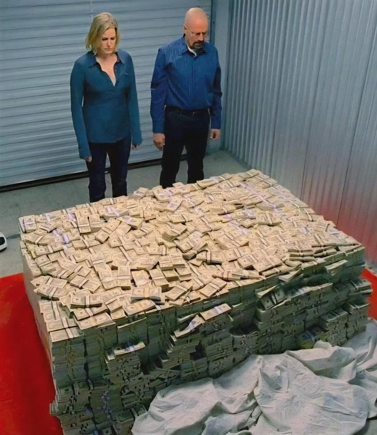 A bald man with a goatee in a blue button-up shirt and black pants stands beside a blonde woman in a blue long-sleeve shirt and jeans. Both look down at a massive rectangular cube of tightly stacked and banded US dollar bills filling most of the foreground. They stand inside a storage unit with corrugated metal walls; a red floor section and a crumpled white sheet are visible near the base of the cash pile.