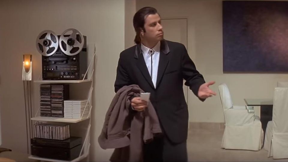 A man in a black suit, white shirt, and black bolo tie stands in a beige living room, holding a brown jacket and a small white cup. One hand is raised, palm up, and he looks off to the side with a blank, puzzled face. To his left, a white shelf holds a reel-to-reel tape deck, stacked discs, and audio gear. White slipcovered chairs and a dark painting appear on the right.
