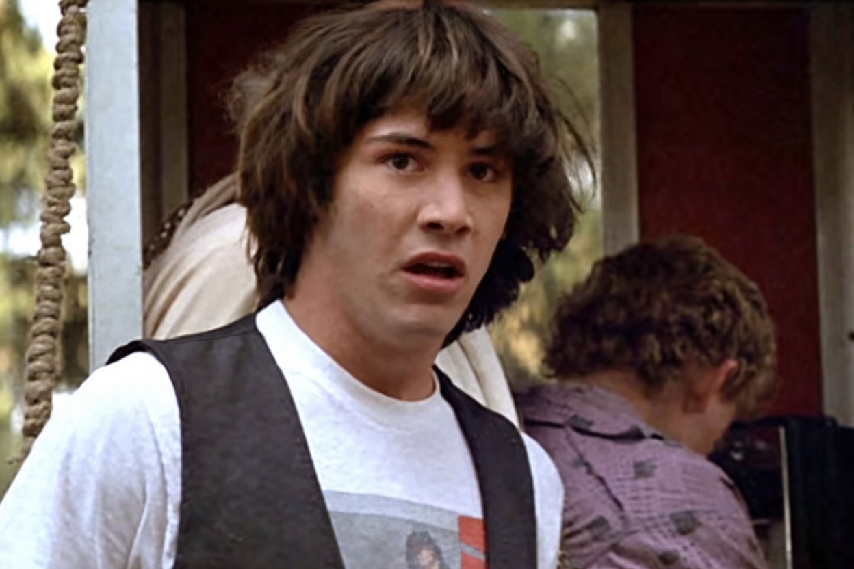 A young man with shaggy medium-length brown hair and bangs, wearing a white graphic t-shirt under a dark vest, stares off-camera with wide eyes and a slightly open mouth in a startled or stunned expression. Behind him are blurred outdoor elements, a red wall/door area, rope, and other people partially visible.