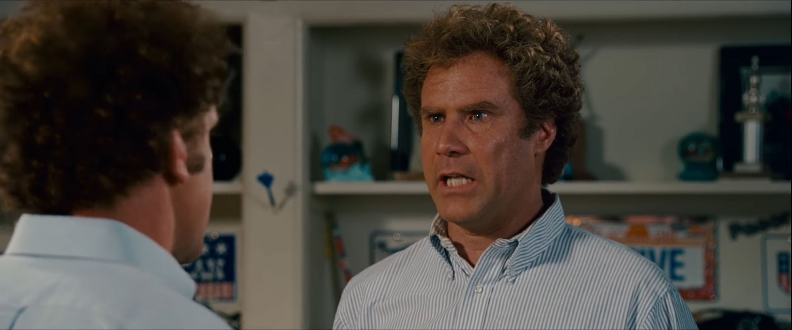 A film still shows two men indoors. On the right, a man with curly light-brown hair and a blue-and-white striped button-down shirt faces left, mouth open mid-sentence, eyes wide and intense. On the left, the back of another man’s dark curly head and light collared shirt fills the foreground, out of focus. Behind them are white built-in shelves holding trophies, decorative objects, and license plates.