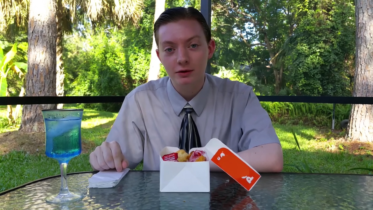 A young man with slicked-back hair sits at an outdoor glass table, wearing a light gray button-down shirt and a black-and-white striped tie. He faces the camera with a flat, unimpressed expression and slightly open mouth. On the table are a tall glass of bright blue iced drink, a folded white napkin under his hand, and an open white fast-food box containing a few chicken nuggets and sauce packets, with an orange Popeyes lid propped open beside it. Behind him is a sunny yard with grass, trees, and a black railing.