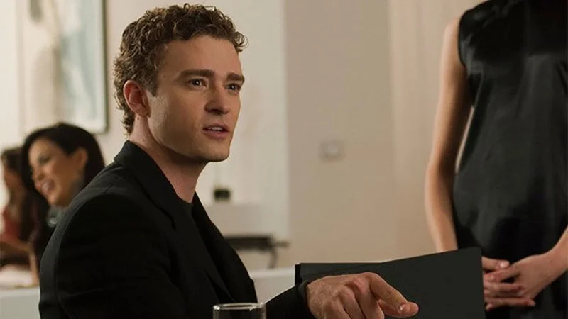 A man with curly light-brown hair wearing a black suit jacket over a black shirt sits at a table, pointing forward with his right index finger while his left hand rests on a black folder or menu. His mouth is slightly open as if speaking. In the background a woman in a sleeveless black dress stands with hands clasped, and other people are seated and blurred at a nearby table in a bright indoor space.