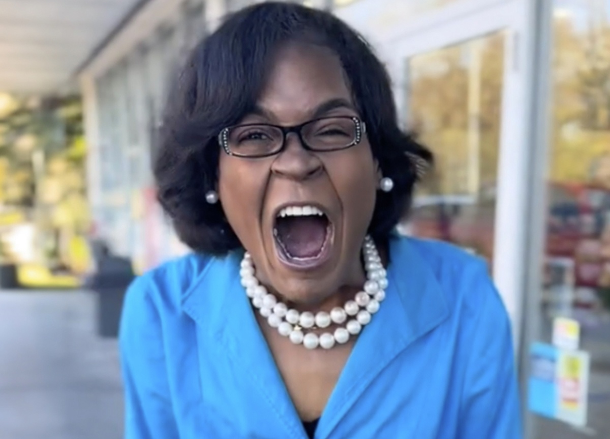 Close-up of a Black woman with a short dark bob, black-rimmed glasses with sparkly temples, pearl earrings, and a multi-strand pearl necklace over a bright blue collared top. Her mouth is stretched wide open in an extreme yell or scream, teeth and tongue visible, eyes narrowed. Background is an outdoor covered walkway beside glass storefront doors and autumn trees.