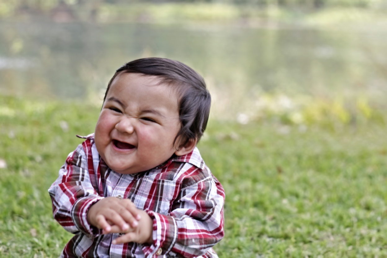 A young toddler with short dark hair sits on green grass outdoors near a body of water. The child wears a red, white, and blue plaid long-sleeve shirt, has hands clasped together in front, eyes squinted shut, and mouth wide open in a big laughing expression.