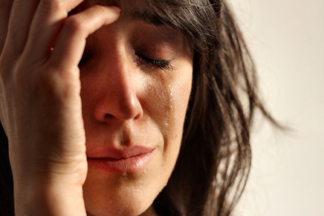 Close-up photo of a dark-haired woman with eyes closed, a visible tear on her cheek, lips slightly downturned, and one hand pressed to her forehead against a plain light background.