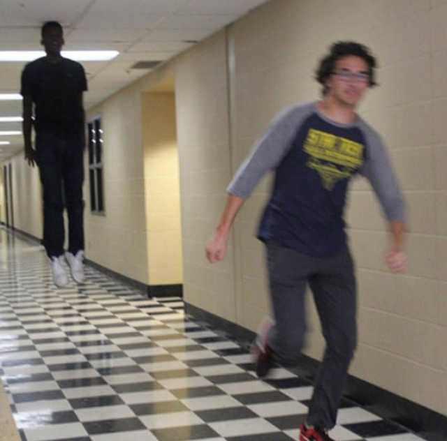 A school hallway with beige cinder-block walls, fluorescent ceiling lights, and a black-and-white checkered floor stretching into the distance. In the right foreground, a young man in a navy-and-gray shirt, gray pants, and red shoes runs toward the camera, motion-blurred, looking back over his shoulder. Farther down the hall on the left, another young man in a black shirt, dark pants, and white shoes stands upright with both feet clearly above the floor, appearing to float.