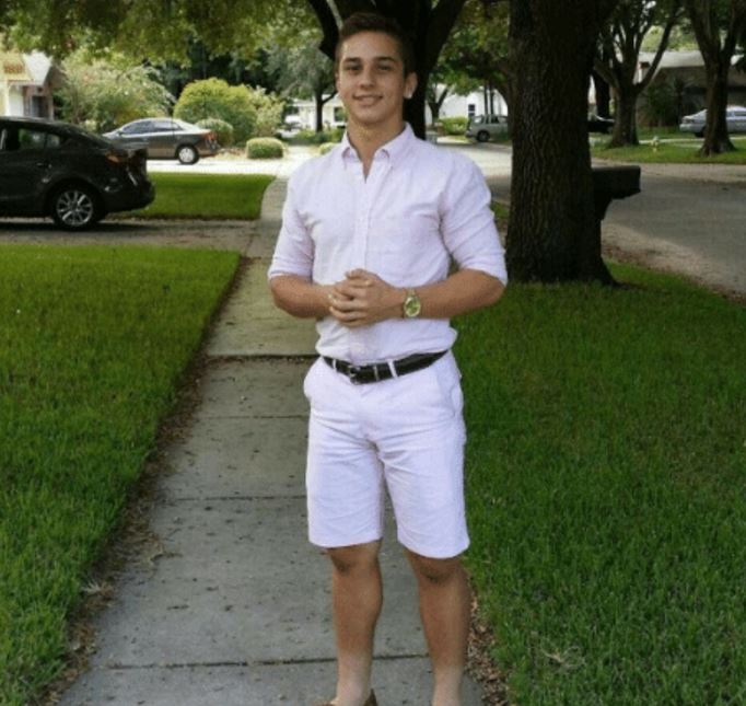 A young man with short brown hair stands facing the camera on a concrete sidewalk, smiling with his hands clasped in front of him. He wears a light-colored short-sleeve button-up shirt with sleeves rolled, white shorts, a black belt, a gold watch, and brown shoes. The background shows a suburban street with large trees, green lawns, parked cars including a black sedan and a gray SUV, houses, and a person sitting on a bench.