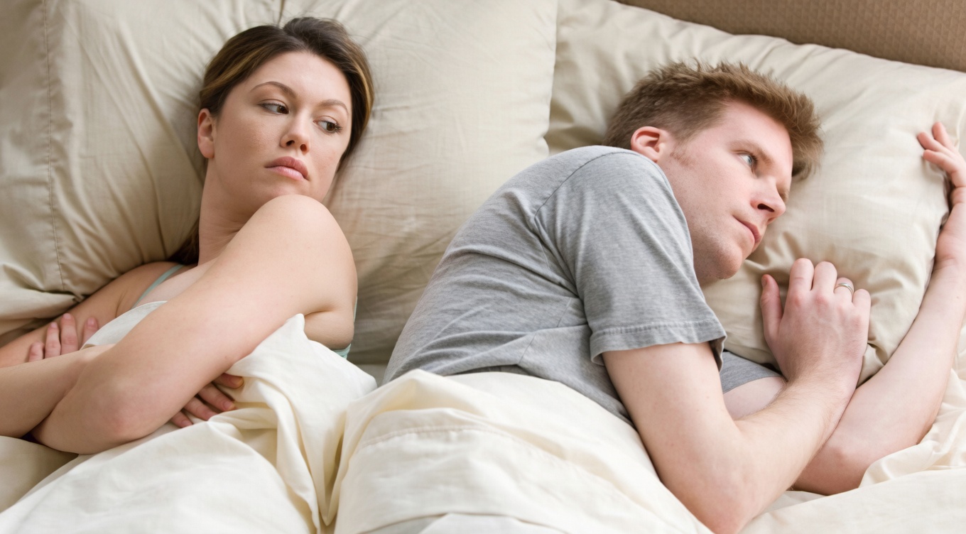 A man and a woman lie in bed under cream sheets and pillows. The woman (left) is on her side facing the man, eyes open with a concerned or suspicious expression. The man (right) lies on his side facing away from her, eyes open with a distant, thoughtful look, one hand near his face.