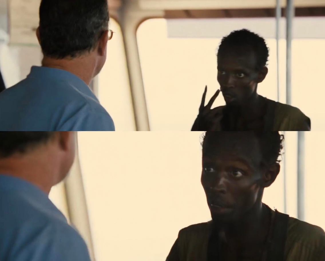 Two stacked film stills. Top: a man in a blue shirt seen from behind faces a thin dark-skinned man who holds up two fingers near his face with an intense stare. Bottom: closer shot of the same thin man looking sideways at the man in blue, mouth slightly open mid-speech, serious expression. Bright window light behind them.