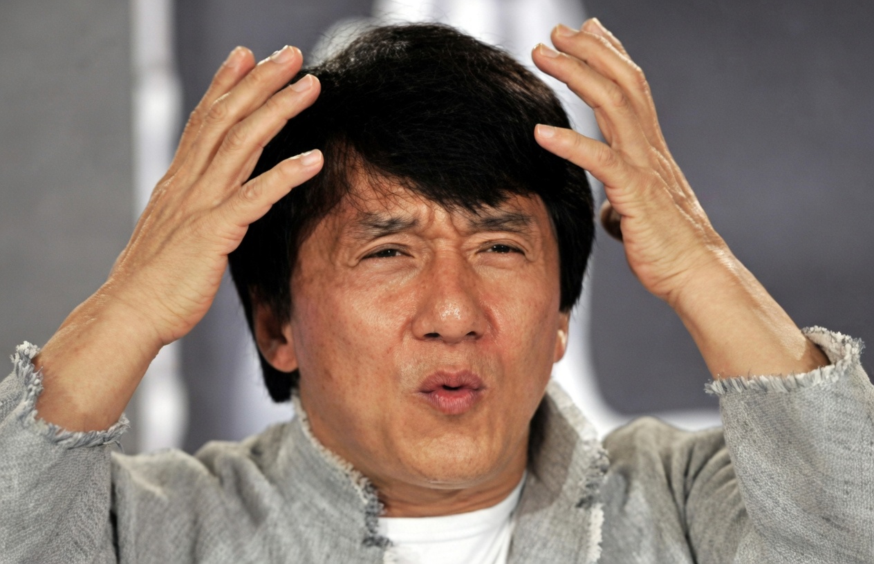 Close-up of Jackie Chan in a gray, frayed-edge jacket over a white shirt. Both hands are raised beside his head with fingers spread. His brows are furrowed, his eyes are narrowed, and his lips are pursed. The background is a soft gray blur. No text is printed on the image.