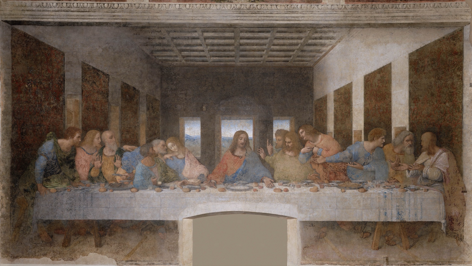 A wide mural of thirteen people seated along one side of a long table in a large room. The central figure wears red and blue robes, has long hair and a beard, and sits with both arms extended on the table. Six figures sit to the left and six to the right, leaning, pointing, turning, and gesturing toward one another. The table has a white cloth, plates, glasses, bread, and food. Behind them is a dim interior with three rear windows and a receding coffered ceiling. Patterned wall panels flank the room. An ornamental cornice runs across the top, and a rectangular opening cuts into the wall beneath the table.