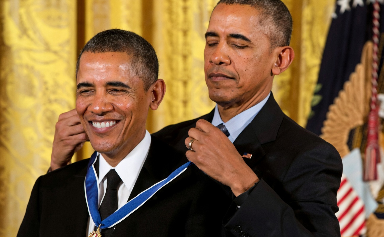 Two identical men in dark suits stand close together in front of gold curtains, with part of an American flag visible at the right. The man on the left smiles broadly while wearing a medal on a blue ribbon with white edges. The man on the right has his eyes closed and is using one hand to place that ribbon around the other man's neck. No text is printed on the image.