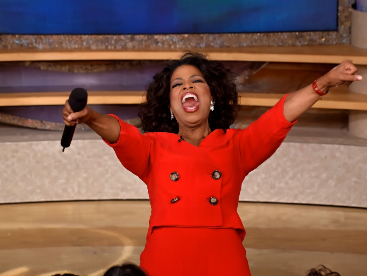 A woman with curly dark hair wearing a bright red double-breasted jacket and skirt stands center stage with both arms raised high and wide, mouth open in a shout or cheer, holding a black microphone in her right hand. A red bracelet is on her left wrist. The background shows a brightly lit studio set with gold curtains and blue lighting. Audience heads are partially visible at the bottom.