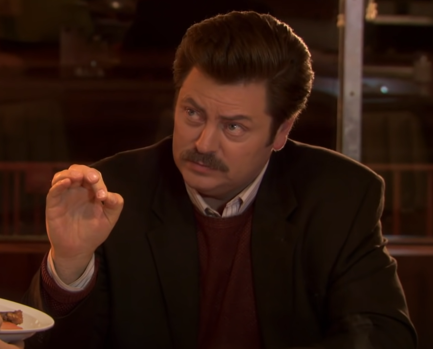 A man with tall swept-back brown hair and a thick mustache sits indoors in warm light, wearing a dark blazer over a burgundy sweater and a white collared shirt. He looks off to the side with a furrowed brow and a tight, unimpressed mouth. One hand is raised, thumb and forefinger pinched together as if inspecting something tiny. A plate of food is partly visible at the lower left; the background is a dim restaurant or bar interior.