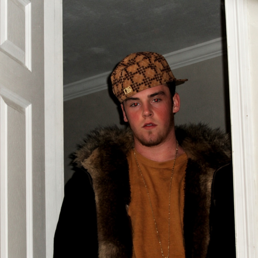 A young man stands in a white doorframe looking toward the camera. He wears a brown checkered flat cap tilted slightly, a mustard-yellow T-shirt, a thin gold chain, and a dark jacket with a thick brown fur-lined hood. He has short facial hair and a neutral, slightly open-mouthed expression; red-eye from the flash is visible. The background is a plain indoor wall and ceiling.
