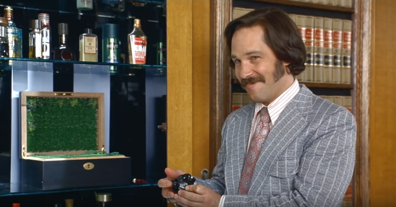 A smiling man with shoulder-length brown hair and a thick mustache wears a gray patterned suit, striped shirt, and patterned tie while holding a small black bottle in both hands. To his left, a wooden box sits open, showing a bright green interior. Behind him is a glass cabinet of assorted bottles, including labels reading BRUT and MUSK, and a bookshelf of matching bound volumes against wood paneling.