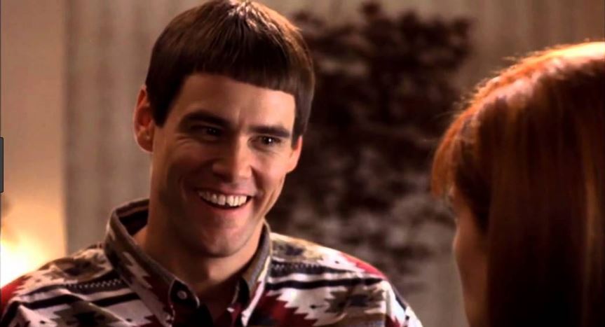 A man with a straight dark bowl cut and a red, black, and white geometric sweater grins widely, teeth showing, while looking at a red-haired person seen from behind on the right. The background is a soft, out-of-focus indoor room. No text is printed on the image.