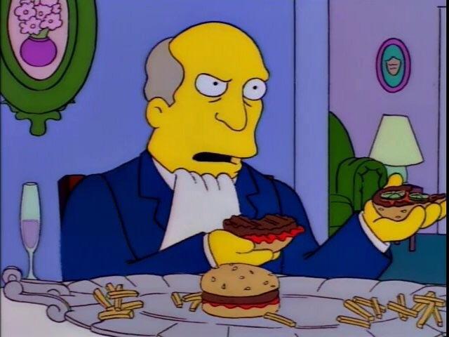 A bald, yellow-skinned cartoon man in a dark blue suit and white shirt sits at a dining table with wide eyes and an open mouth, looking unconvinced. He holds a dark grilled sandwich in one hand and a lettuce-topped burger in the other. On the white tablecloth are a sesame-seed hamburger and scattered fries, with a stemmed glass to the left. The room has purple walls, a green oval frame with pink flowers, a small round portrait, a lamp, and a green chair.