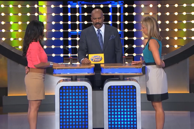 A game-show stage with a wall of round white lights and blue-green panels. A bald man with a mustache, in a gray suit and blue tie, stands center looking down at a yellow sign that reads Family Feud. Two contestants face inward at matching blue podiums with buzzers: on the left, a woman with long dark hair in a pink top and tan skirt, one hand on her hip; on the right, a blonde woman in a teal top and a white-and-black skirt. No caption text is printed on the image.
