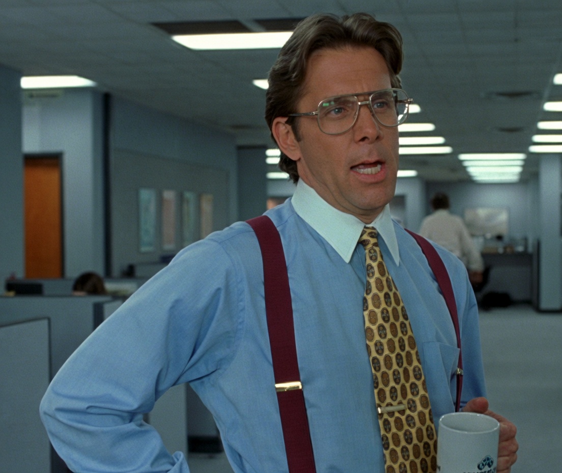 A man with brown hair and large glasses stands in a bright office cubicle area. He wears a light blue dress shirt, red suspenders, and a yellow patterned tie, holding a white coffee mug in one hand with the other hand on his hip. His mouth is slightly open as if speaking. Fluorescent ceiling lights and office partitions fill the background, with another person visible farther back.