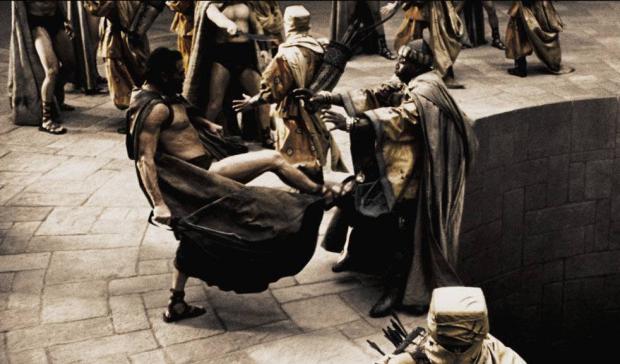 A high-angle shot of a stone courtyard. A muscular, shirtless man in a dark cape and sandals is frozen mid-kick, foot driven into a robed, helmeted man who is falling backward toward a dark pit beside a stone wall. Other cloaked, helmeted figures with spears stand around the pavement. In the lower right, a hooded figure with a quiver looks up. The image is desaturated, bronze-sepia, with motion in the cape.