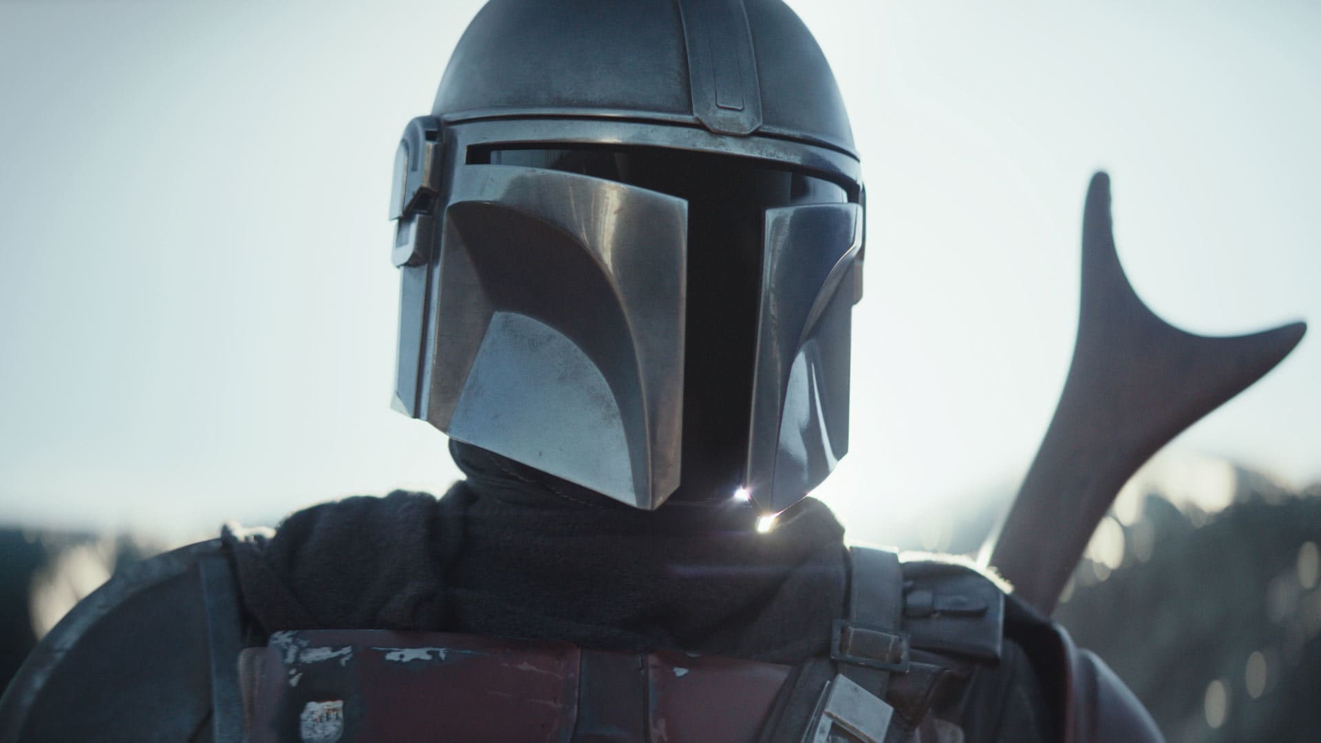 A close-up of a figure in a scratched silver-gray metal helmet with a black T-shaped visor, a dark fabric cowl, and worn reddish-brown chest armor with straps. The background is a bright, hazy sky. A dark curved silhouette rises behind the figure’s right shoulder, and a small bright glint sits near the chin.