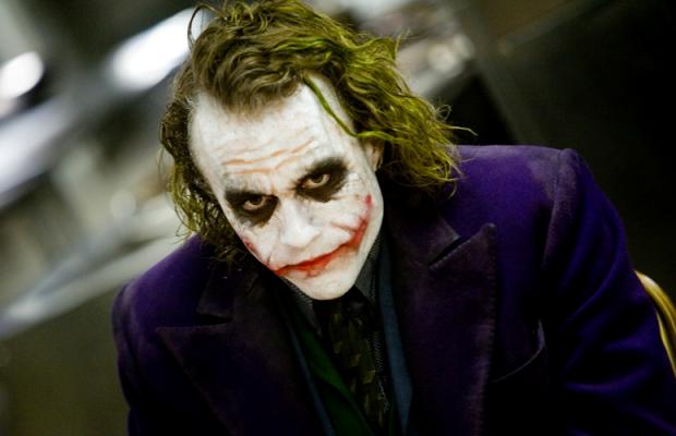 A person with messy green hair leans in toward the camera. White face paint covers the face, with heavy black makeup around the eyes and a red smile smeared past the corners of the mouth. They wear a purple coat over a dark green vest, a patterned shirt, and a dark tie. The background is a blurred indoor space with bright overhead lights. The head is tilted and the stare is direct.