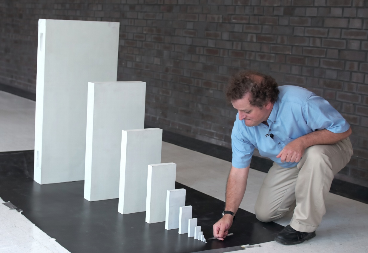 A man in a light blue shirt and khaki pants kneels on a black floor mat beside a row of upright white rectangular blocks. The blocks decrease sharply in size from a very tall slab on the left to a tiny piece on the right; he is touching or about to tip the smallest one with his finger. A dark brick wall is in the background.