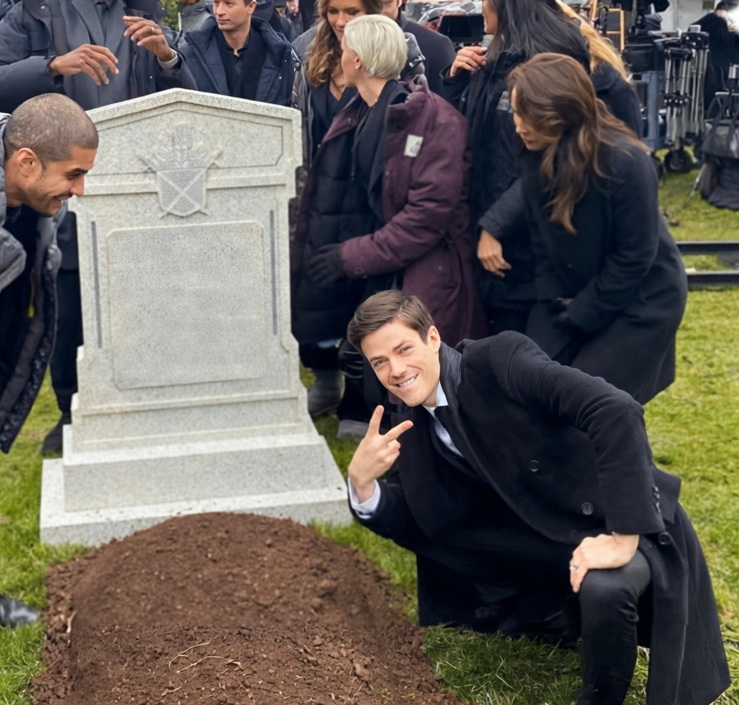 A man in a dark coat crouches on grass beside a fresh mound of brown dirt and a tall gray headstone with a blank rectangular plaque and a crest. He smiles at the camera and holds up a peace sign. Behind him, several people in coats stand near film/camera equipment, looking toward the headstone or each other on what appears to be a set.