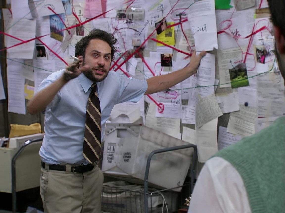 A wild-eyed man with dark curly hair and a beard, wearing a light blue short-sleeve dress shirt, striped brown tie, and khaki pants, stands in a cluttered office. He holds a cigarette in one hand and presses the other against a wall densely covered in papers, photos, notes, and red string connecting them in a chaotic web. He faces partly toward a person in the foreground (seen from behind in a green sweater vest) while gesturing intensely. The room has filing cabinets, mail bins, and scattered documents.