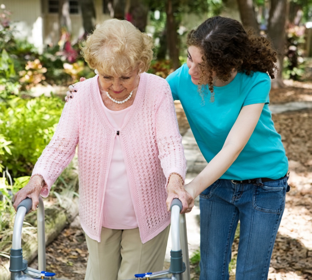 An elderly woman with short blonde curly hair, pearl necklace, pink open-knit cardigan, and light pants walks with a silver walker on an outdoor path. A younger woman with dark curly hair in a bright teal t-shirt and blue jeans stands beside her, one hand on the elder’s shoulder and the other on the walker, leaning in with a concerned expression. Green plants, trees, and a house are in the background.
