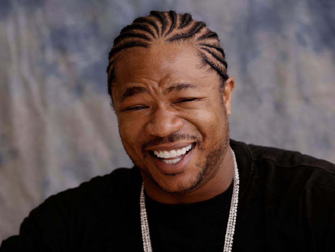 Close-up portrait of a smiling man with cornrow braids swept back, light facial hair, wearing a black shirt and a multi-strand diamond tennis chain. He is squinting one eye and grinning widely with teeth showing against a mottled gray-blue studio backdrop. No text appears on the image.
