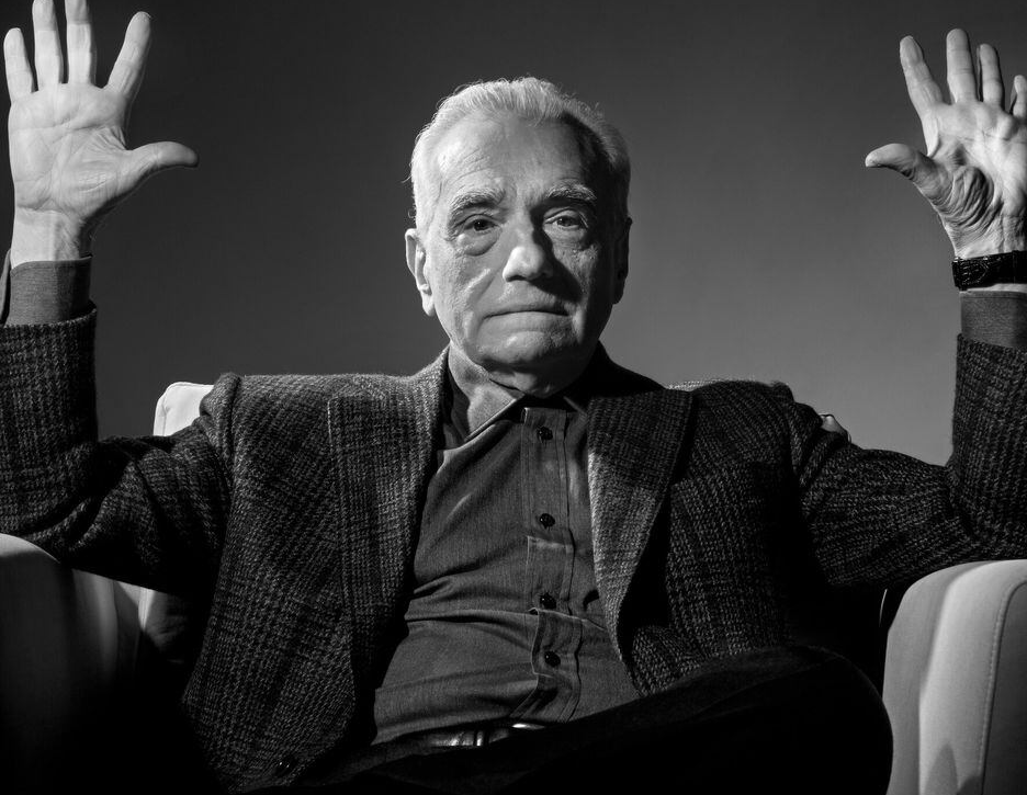 Black-and-white photo of an older man with white hair sitting in a light-colored chair. He wears a textured plaid blazer over a dark button-up shirt. Both arms are raised to head height with open palms facing forward. His expression is serious and direct toward the camera against a plain dark gradient background.