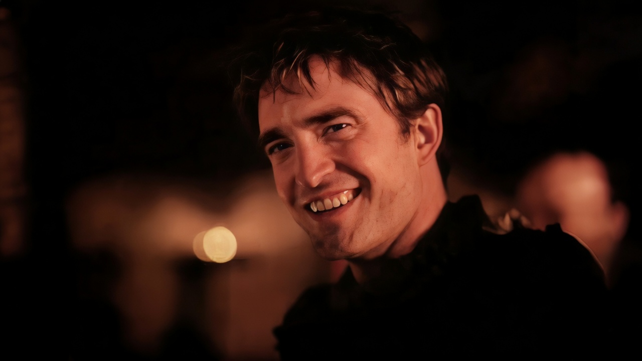 A close-up of a dark-haired man turning to look over his shoulder toward the viewer with a wide, toothy grin. The background is dark with out-of-focus warm lights; he wears dark clothing.