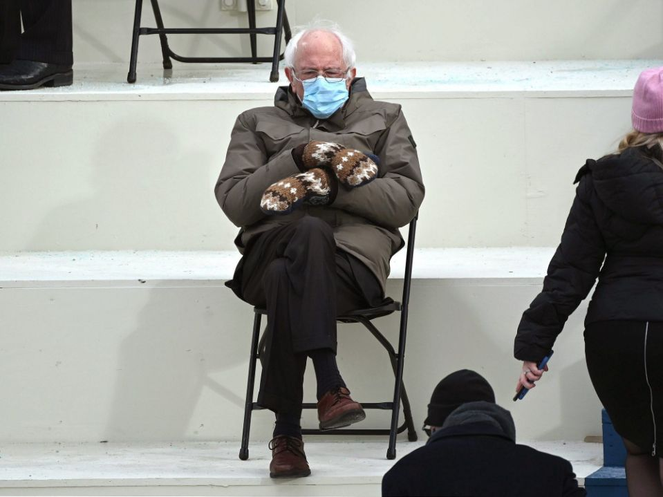 An older man with white hair and glasses sits alone on a black folding chair on wide white steps. He wears a light blue surgical mask, a bulky brown parka, dark trousers, brown shoes, and oversized brown-and-white patterned mittens, with his arms crossed. Partial figures appear at the edges: legs and shoes at the upper left, a person in a pink hat and black coat at the right holding a phone, and the backs of people in dark clothing at the lower right. A metal chair frame sits on the step behind him. The scene is brightly lit and mostly white.