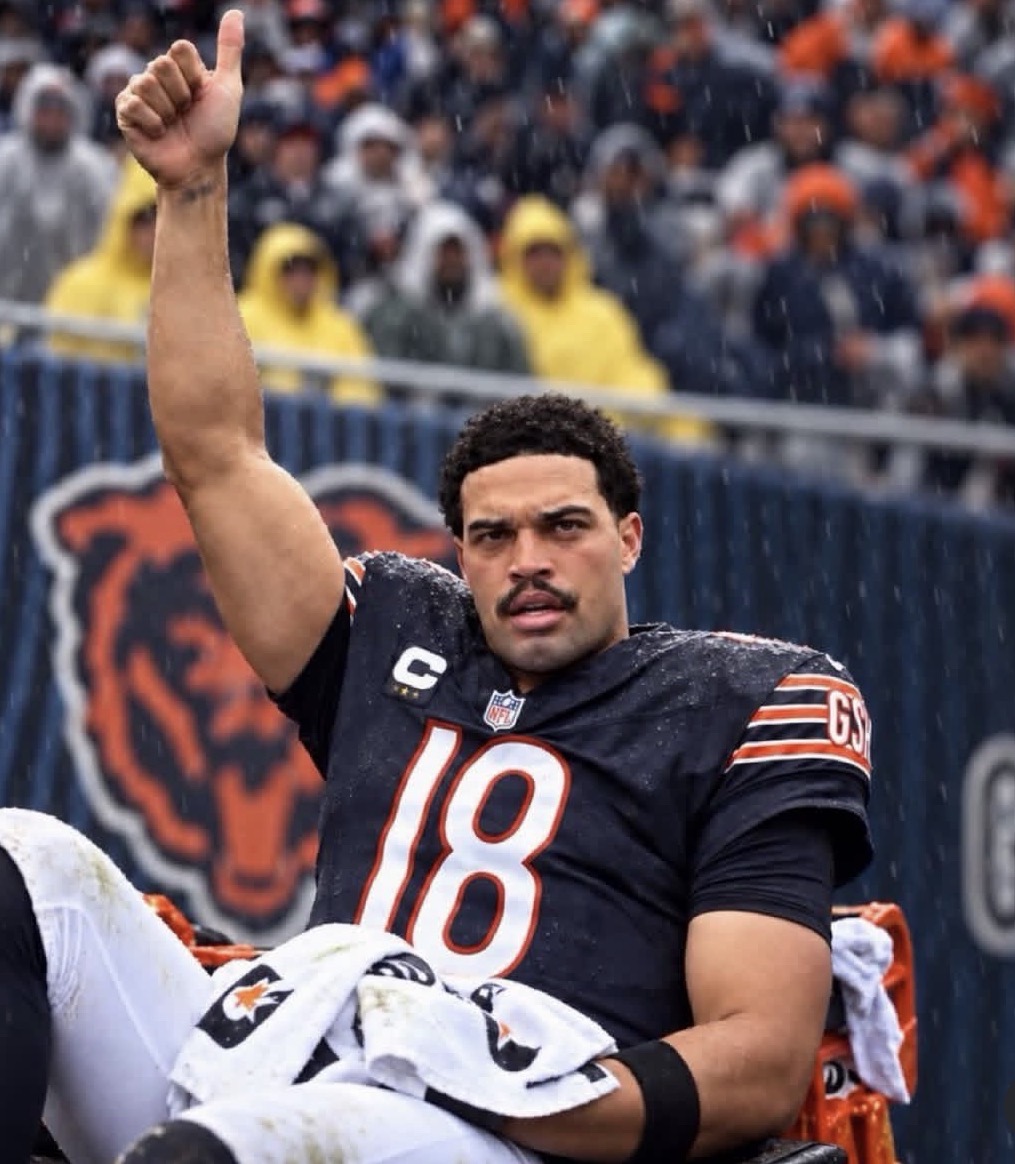 A man with short dark curly hair and a mustache sits on an orange sideline seat in falling rain. He wears a wet dark navy football jersey numbered 18 with a white captain C patch, NFL shield, and GSH on the sleeve, plus grass-stained white pants. His right arm is raised in a thumbs-up. A white towel with logos lies on his lap. Behind him is a stadium crowd in rain ponchos (many yellow) and a large bear-head logo on a blue wall.