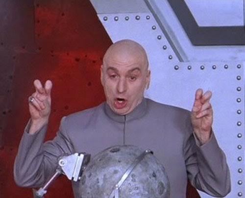 A bald man in a plain gray high-collared jacket faces forward with his mouth open mid-speech. Both hands are raised beside his head, each making an air-quote gesture with the index and middle fingers; a ring shows on one hand. A gray, cratered sphere on a metal clamp sits in the lower foreground. The background is a riveted metal wall, reddish on the left and pale gray with a dark rectangular frame on the right.
