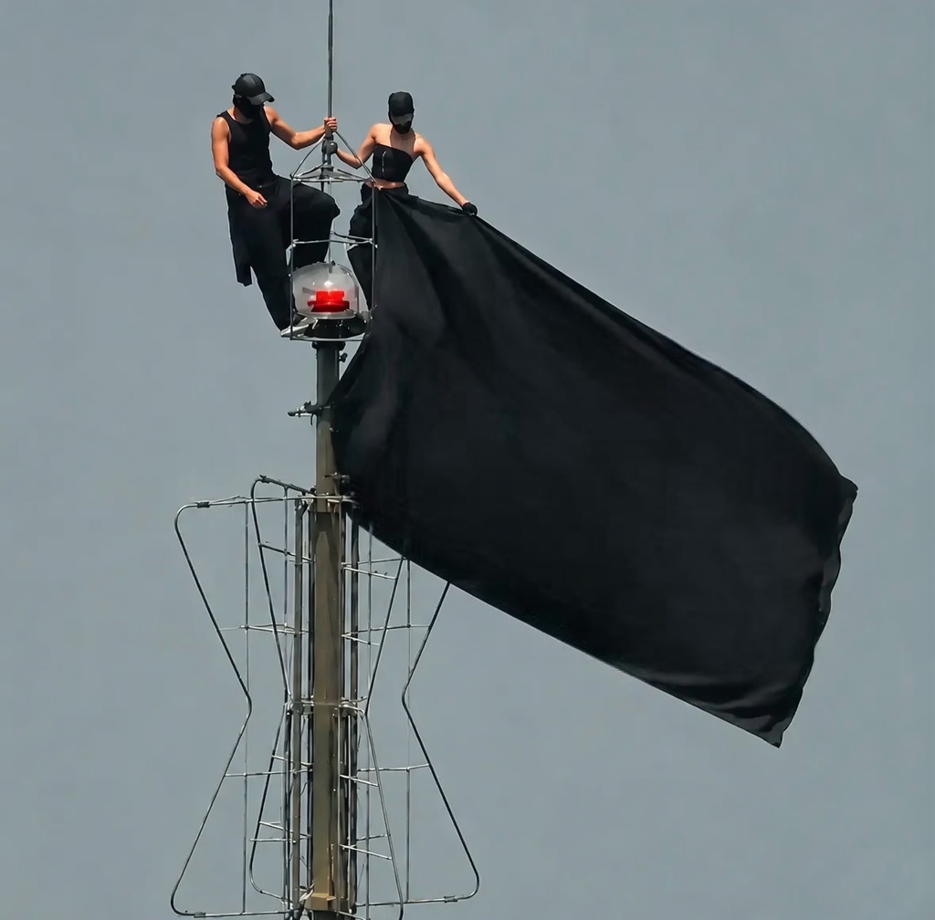 Two people dressed entirely in black (tank tops, pants, caps, face coverings) stand at the very top of a tall metal lattice antenna/tower against a plain gray sky. They are holding and unfurling a large solid black flag that billows dramatically to the right. A red aviation warning light is visible on the tower below them. A small “8/8” mark appears in the upper-right corner.