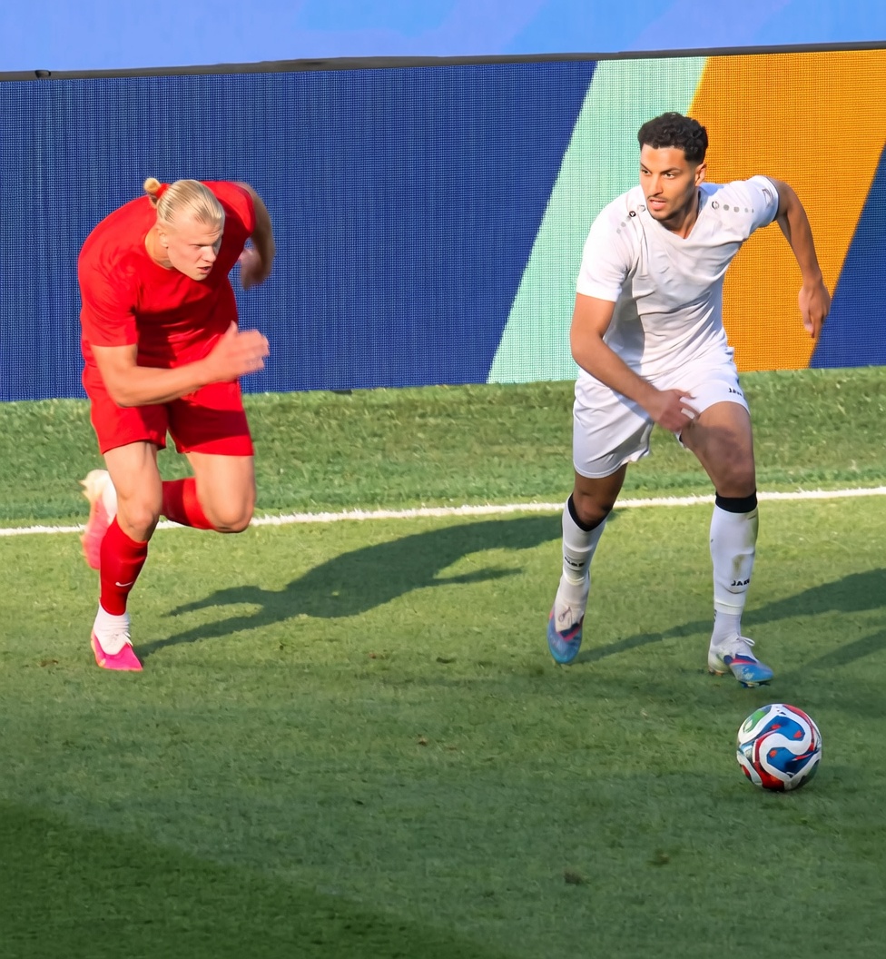 Two male soccer players sprinting on a green grass pitch. On the left, a tall blonde player in a red kit with blue/white trim and the number 9 runs hard toward the right. On the right, a dark-haired player in a white kit with the number 4 runs near a multicolored soccer ball on the ground. A blue LED-style wall is behind them; strong sunlight casts long shadows.
