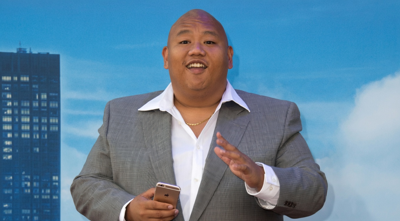 A bald man in a gray suit jacket over an open-collar white dress shirt and thin gold chain stands facing the camera. He holds a gold smartphone in his right hand and gestures outward with his open left hand. His mouth is open in a wide, enthusiastic smile as if mid-explanation. Behind him is a blue sky backdrop with partial “MARVEL STUDIOS” and “SPIDER-MAN” premiere lettering and a skyscraper graphic.
