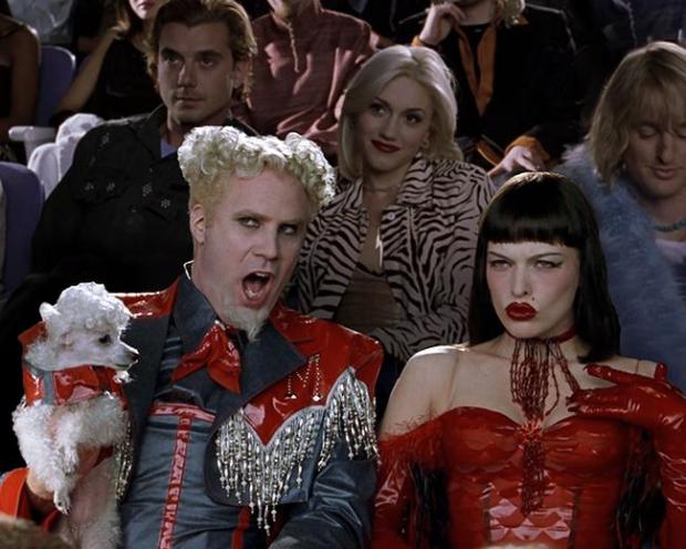A man with short curly platinum-blonde hair and a pale goatee shouts with his mouth wide open. He wears a glossy red jacket with hanging silver fringe over a blue-gray shirt and holds a small white poodle dressed in red. Beside him, a woman with a black bob and blunt bangs, red lipstick, a beaded choker, a shiny red dress, and long red gloves sits with one hand raised. Behind them, audience members are seated in rows, including a man in a dark patterned shirt, a blonde woman in a zebra-print jacket, and a person in a pale blue fur wrap.