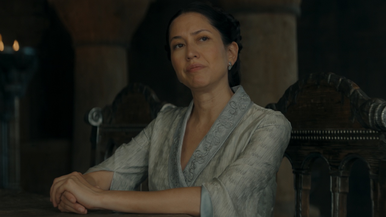 A woman with dark hair in an elegant updo sits at a wooden table in a dim stone hall, wearing a pale gray embroidered robe. Her hands are clasped on the table; she looks upward and slightly aside with pressed lips and a restrained, unimpressed expression. Ornate dark wooden chairs and pillars fill the background.