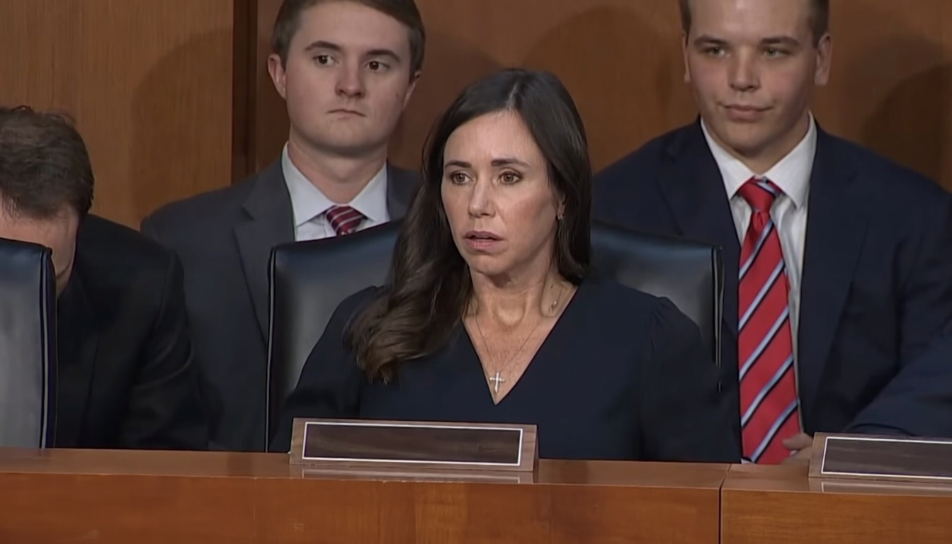 A woman with long dark hair in a navy V-neck top and a small cross necklace sits at a wooden dais, mouth slightly open in a concerned expression. Two young men in dark suits stand behind her (one with a red-and-white striped tie). A man in a suit is partially visible on the far left looking down. Blank wooden nameplates sit on the desk; wood-paneled wall behind.