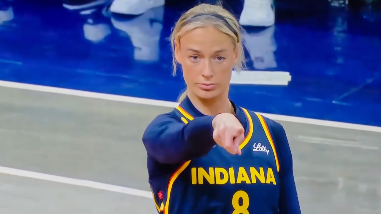 A blonde woman wearing a navy blue Indiana basketball jersey with yellow trim and the number 8 stands on a court, staring intensely forward while pointing her right index finger directly at the viewer. A second hand enters from the lower left, also pointing toward her or the center.