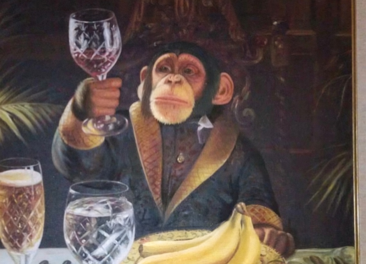 A framed painting of a chimpanzee in a dark jacket with gold-trimmed lapels and cuffs, raising a cut-crystal wine glass of red wine in a toast. It sits at a table with a champagne flute, a water glass, and a bunch of yellow bananas on a plate; dark ornate background with foliage.
